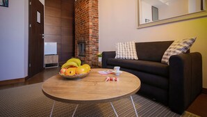 A living room with a brick fireplace, a black sofa with plaid cushions, a round wooden coffee table with fruits and a cup, and a mirror on the wall. - Boogie Aparthouse Old Town (Wroclaw)