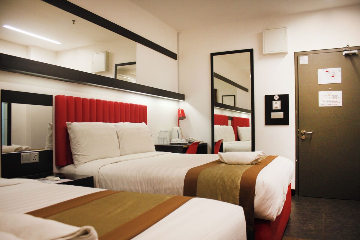 A hotel room with two beds, a red headboard, a large mirror, a door, and a small table with a lamp.