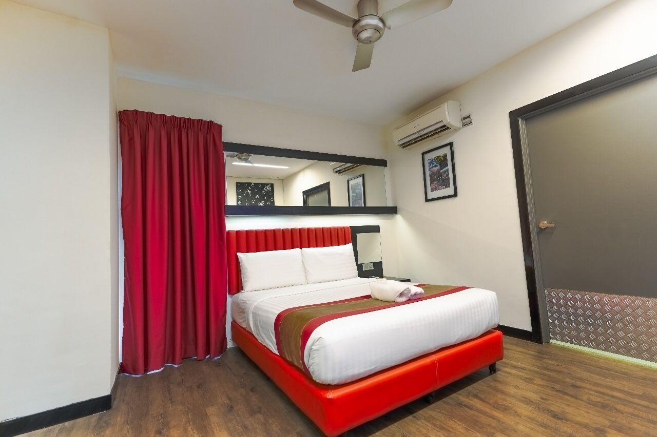 Habitación de hotel con cama, cortinas rojas, ventilador de techo, equipo de aire acondicionado y puerta con felpudo estampado.
