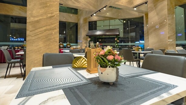 Uma área de refeições moderna com mesas de mármore, um vaso decorativo com flores e vista para a cozinha ao fundo.