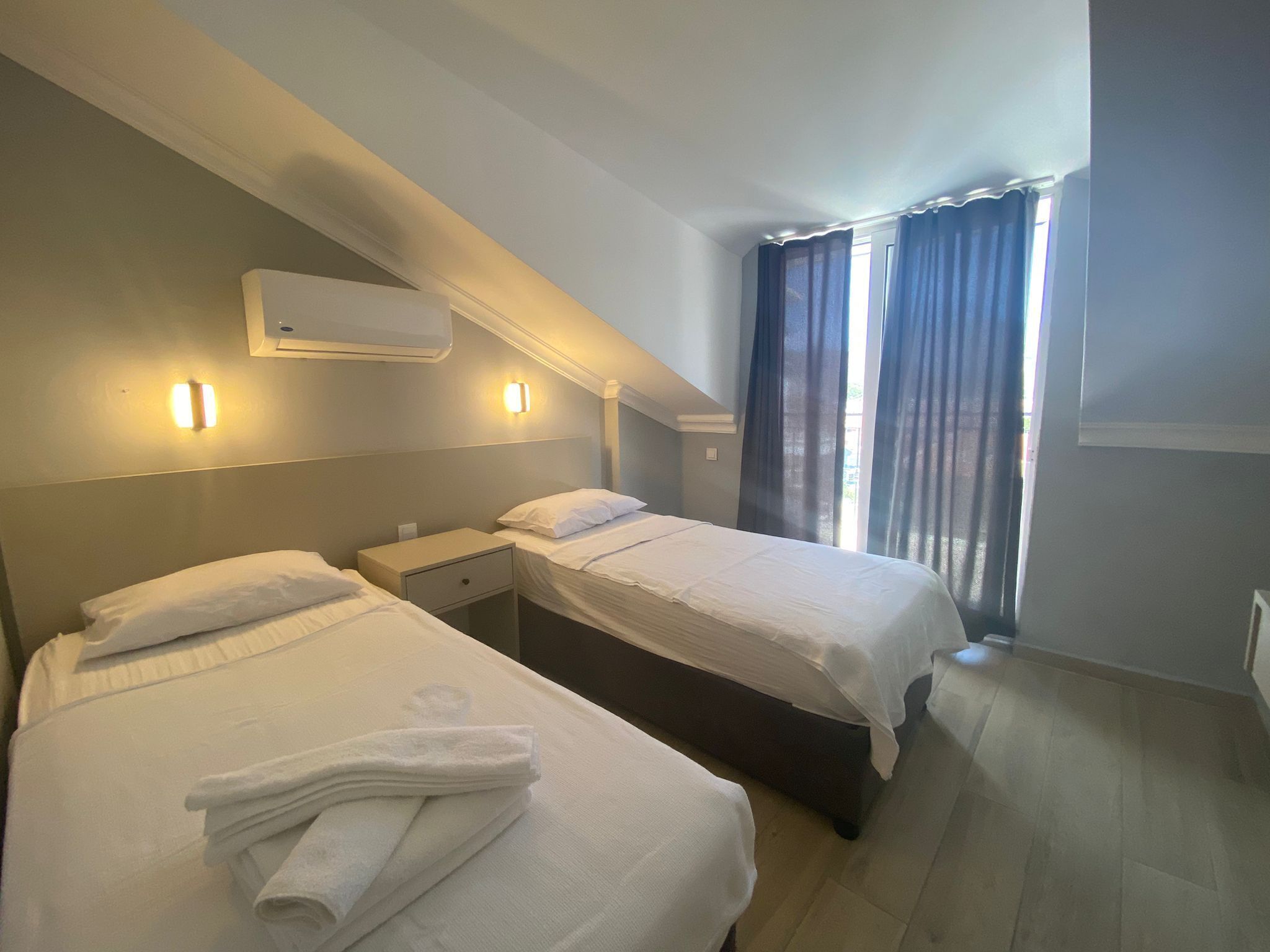 A hotel room with two beds, a ceiling-mounted air conditioner, and a window with curtains.