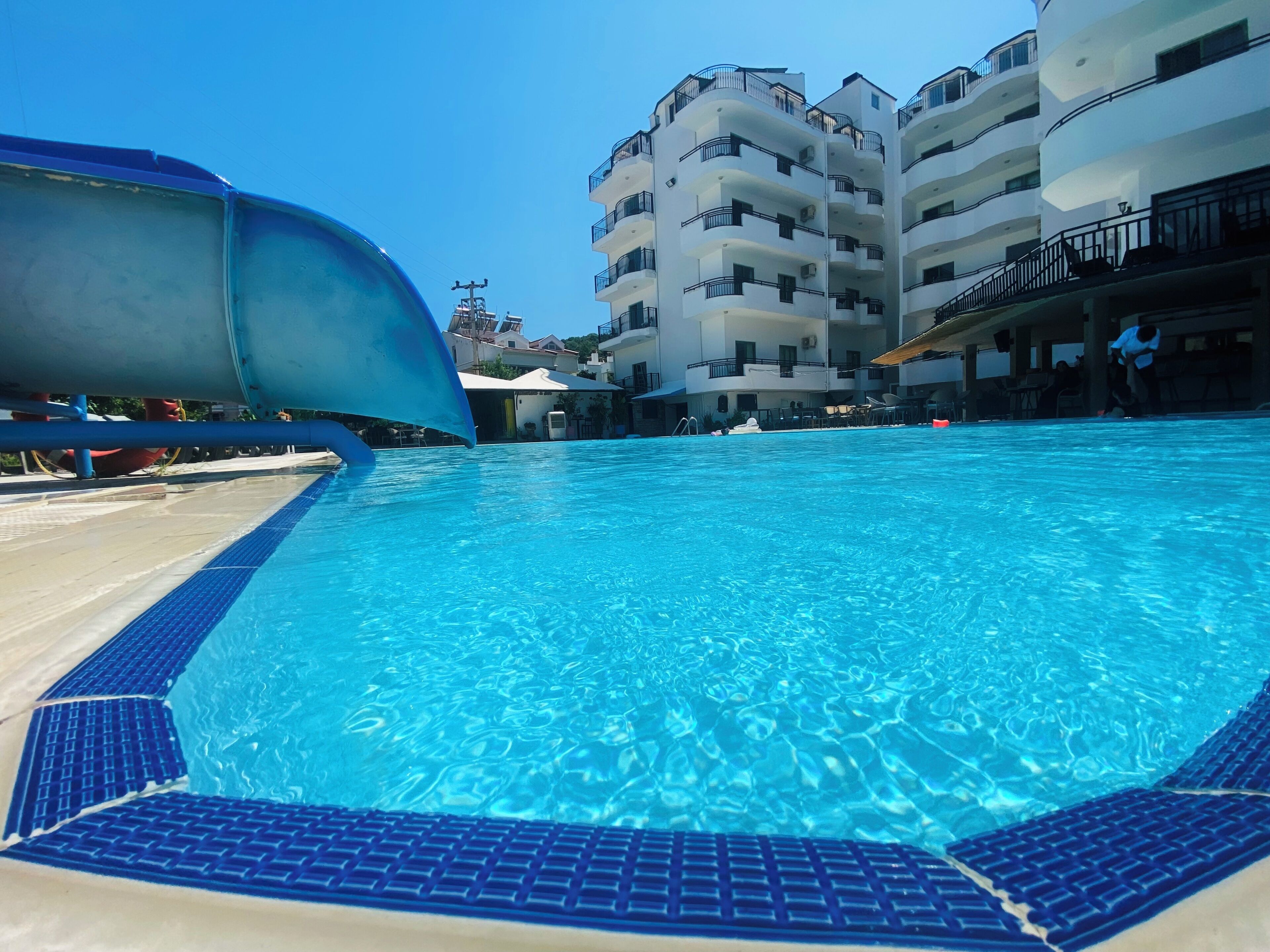 A swimming pool with a blue border, a water slide, and a multi-story building in the background.