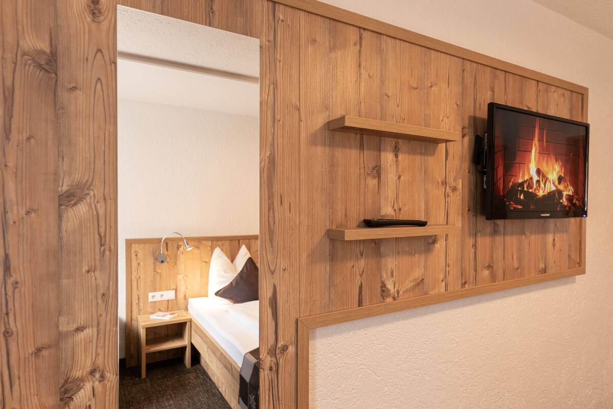 Quarto de hotel com painel de parede em madeira, cama com almofadas, mesa de cabeceira, televisão fixada na parede e lareira decorativa.