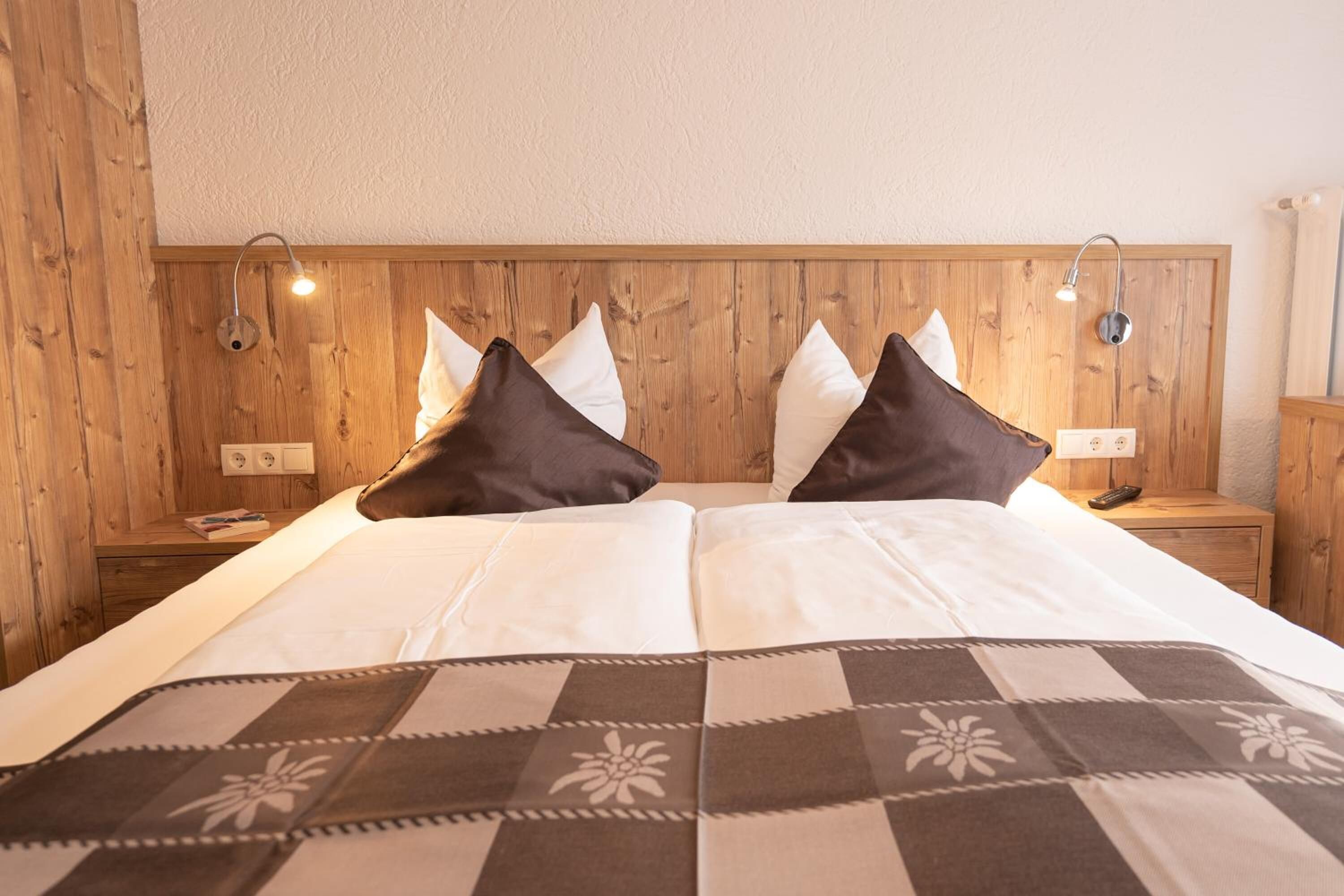 A hotel room with a wooden headboard, two bedside tables, a bed with white bedding and a patterned throw, and two wall-mounted lamps.
