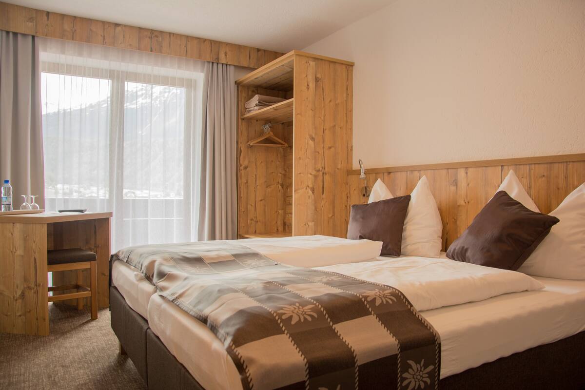 Quarto de hotel com um guarda-roupa de madeira, uma cama com colcha estampada, uma escrivaninha e uma cadeira, e uma janela com cortinas.