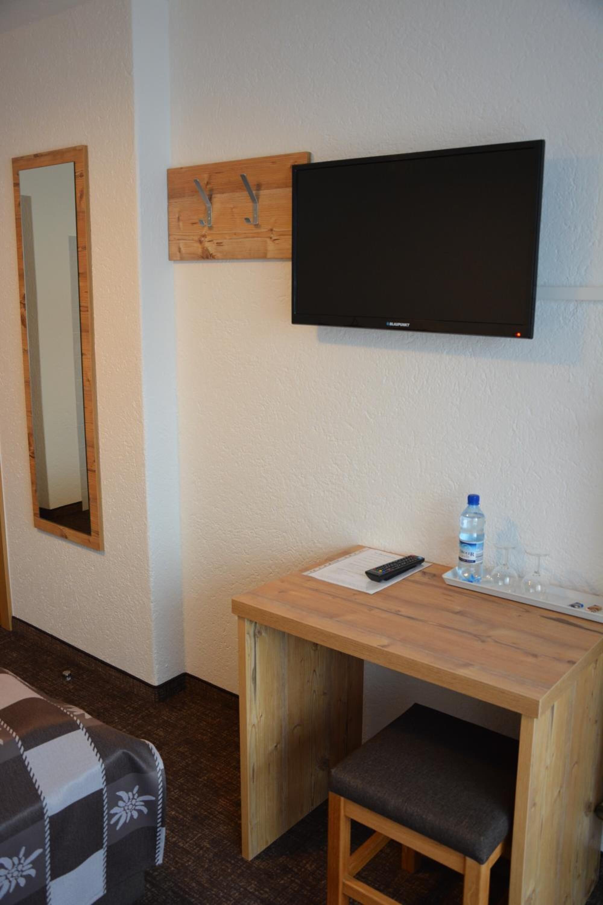 A small room with a wooden desk, a television mounted on the wall, a mirror, a water bottle, and a remote control.