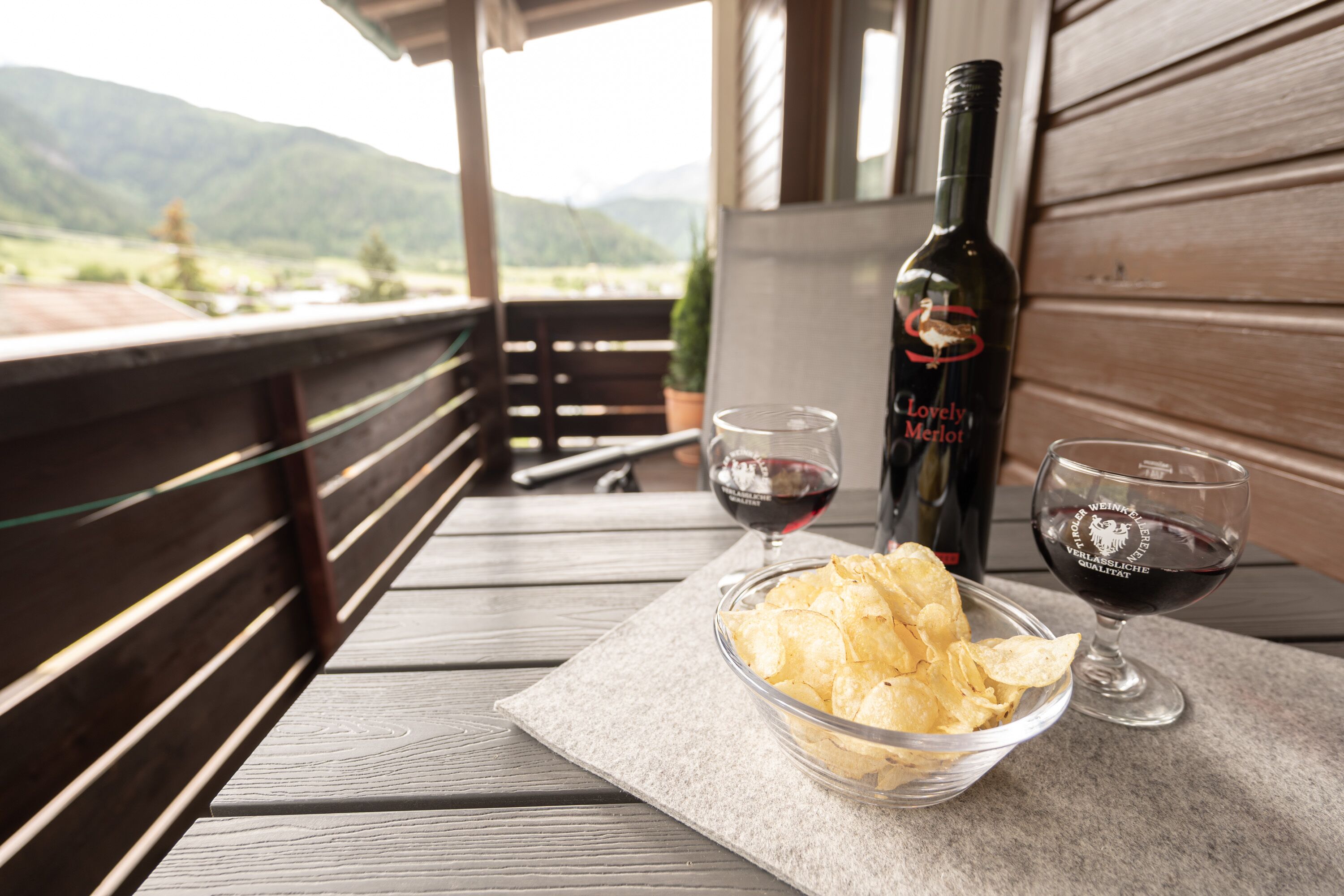 A wooden table with a bowl of chips, two wine glasses, and a bottle of wine on a balcony with a mountain view.