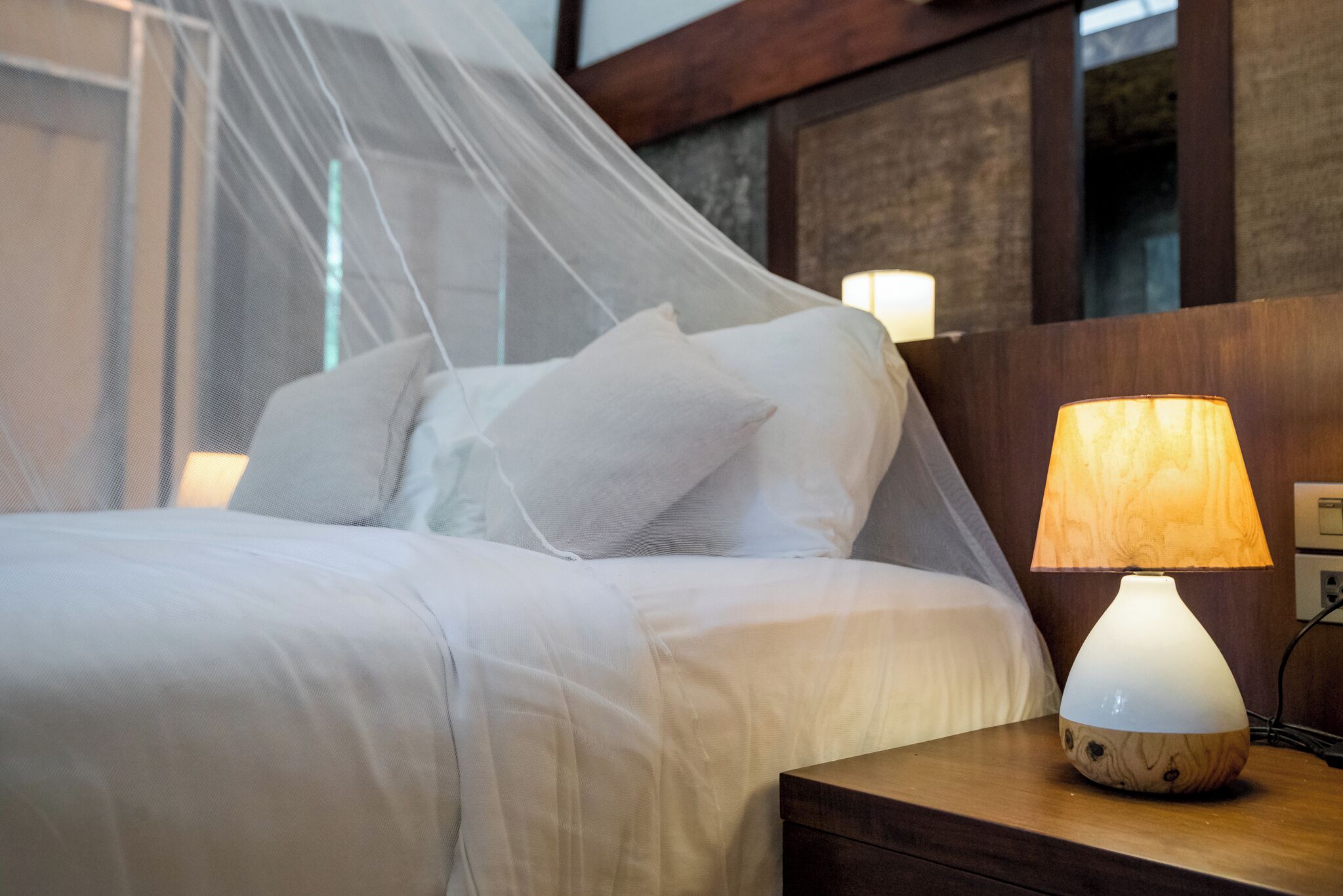 Una cama bien hecha con sábanas blancas, una lámpara de noche y una mesita de madera.