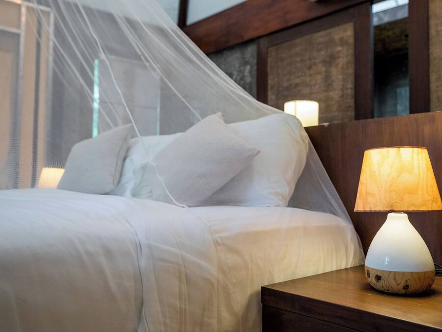 Una cama bien hecha con sábanas blancas, una lámpara de noche y una mesita de madera.