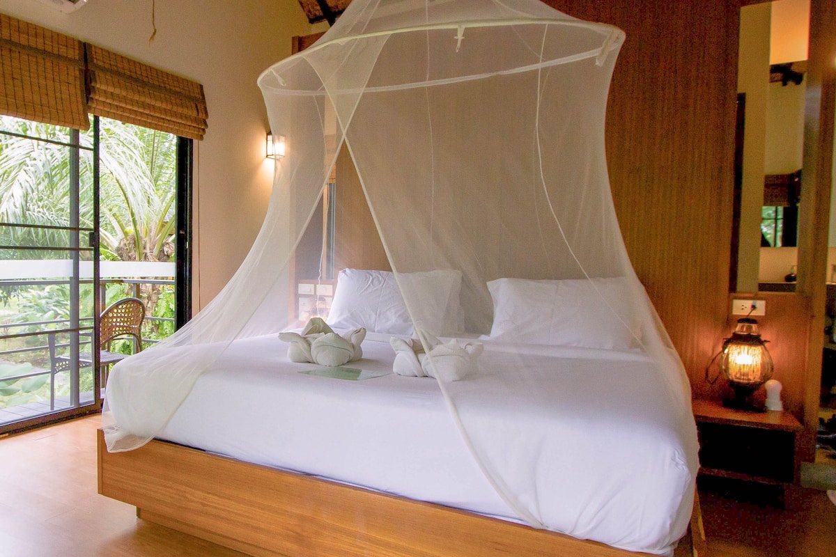 A bed with a canopy, a nightstand with a lamp, a window with a view of greenery, and a ceiling with wooden beams.