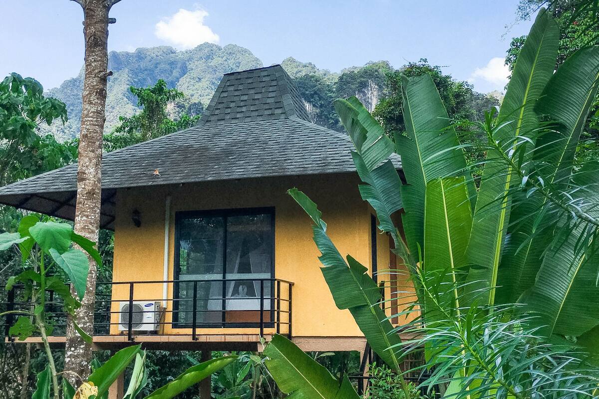 A yellow building with a balcony, surrounded by lush greenery and mountains in the background.