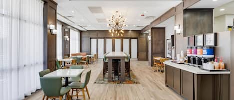 A modern hotel dining area with a central table, seating, a coffee station, and a large window with curtains.