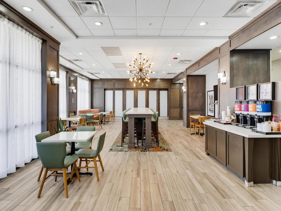 A modern hotel dining area with a central table, seating, a coffee station, and a large window with curtains.
