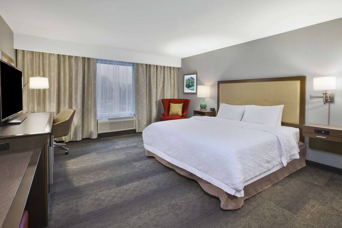 A hotel room with a large bed, a desk, a chair, a TV, and a window with curtains.