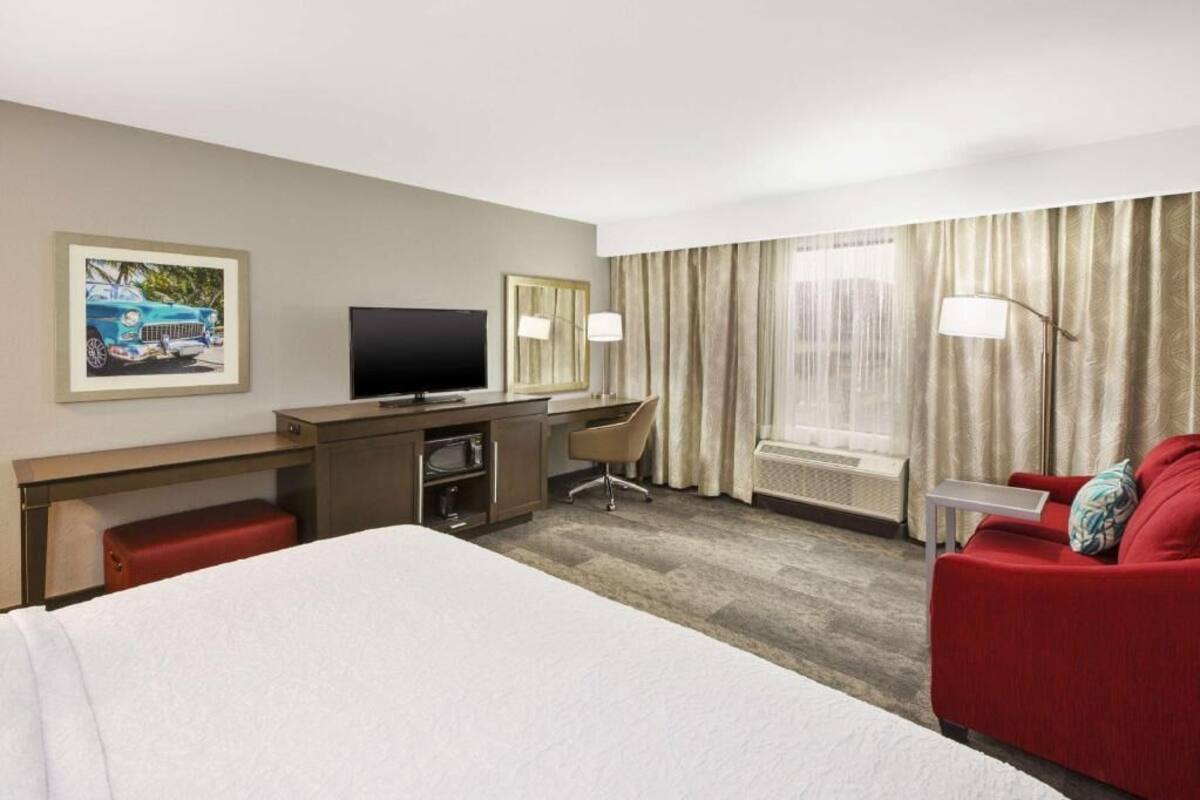 A hotel room with a bed, a desk with a TV, a red sofa, a chair, and a window with curtains.