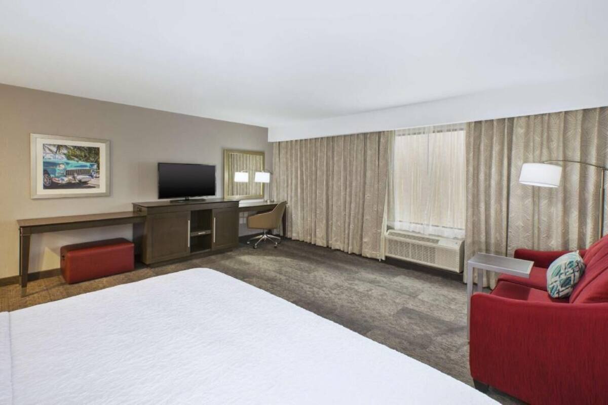 A hotel room with a bed, a desk with a computer, a TV, a red sofa, and a window with curtains.