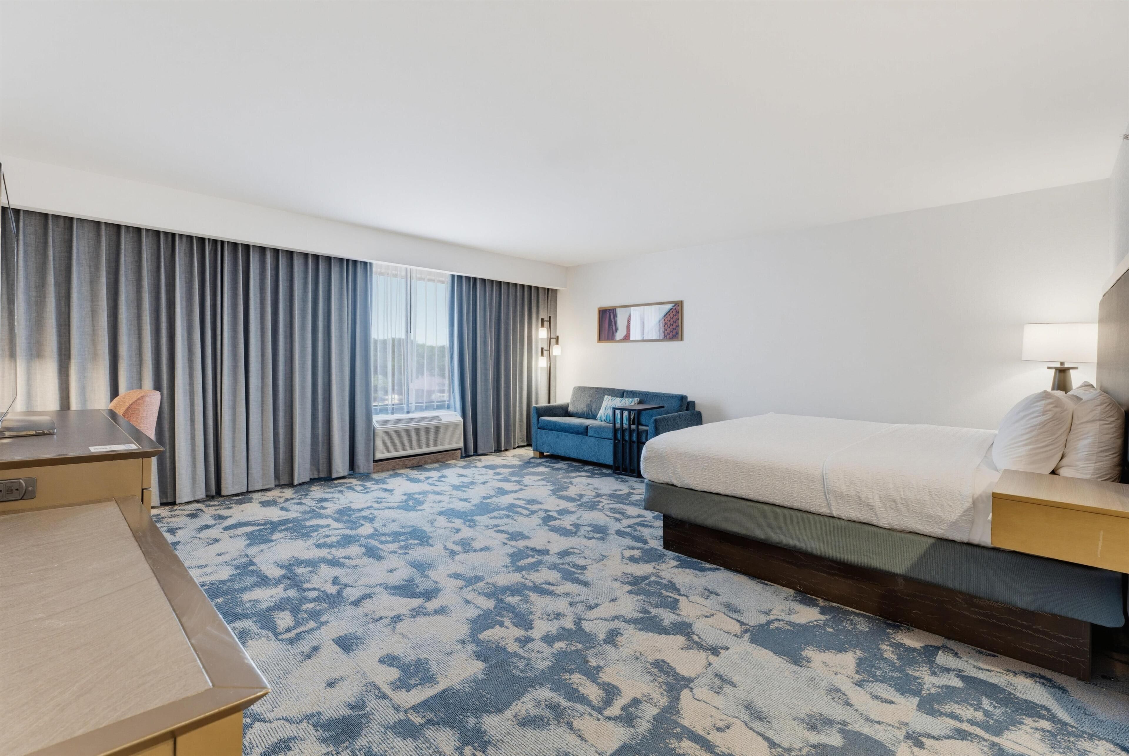 A hotel room with a bed, a desk, a chair, a sofa, and a large window with curtains.