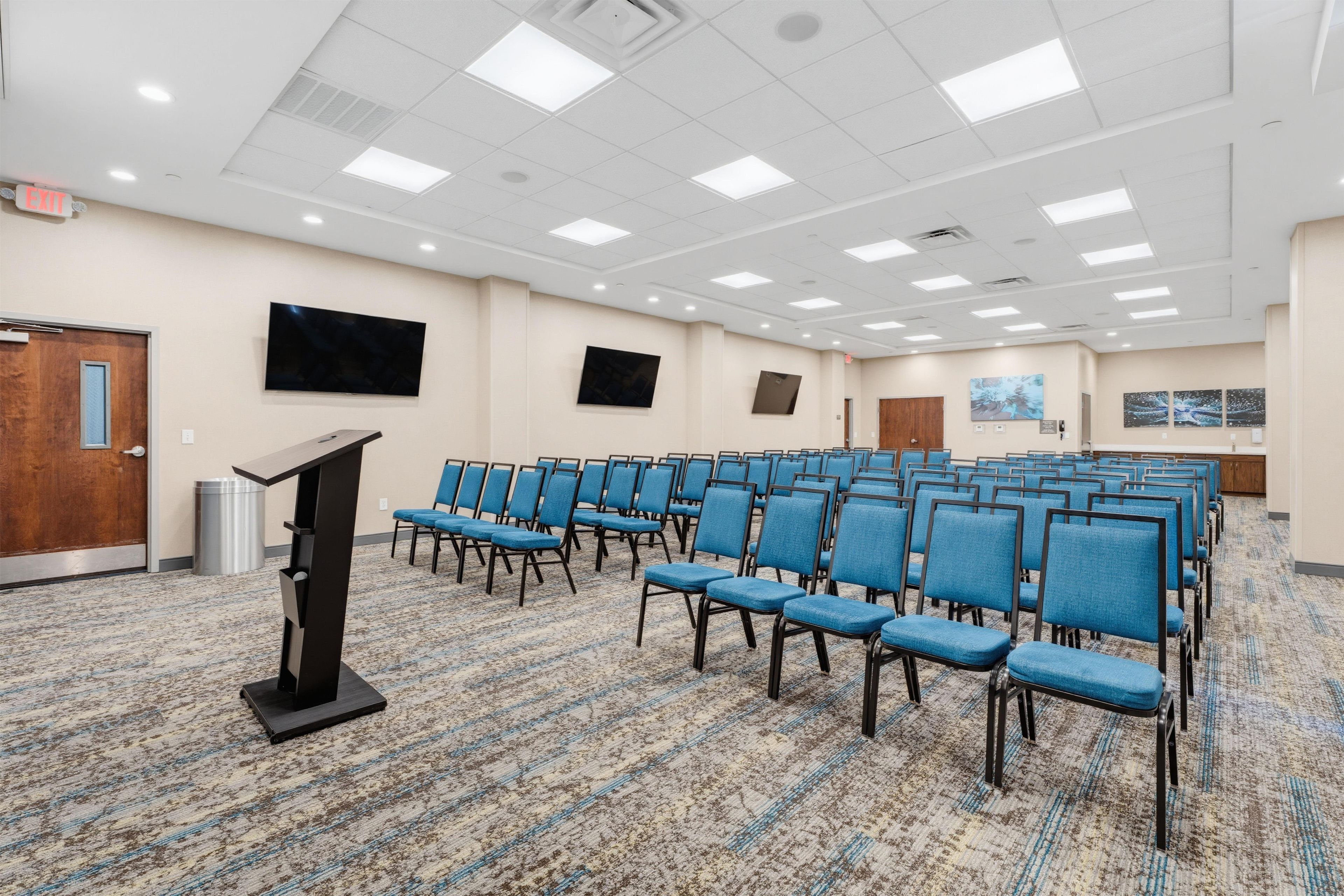 A conference room with rows of blue chairs, a podium, and a flat-screen TV on the wall.