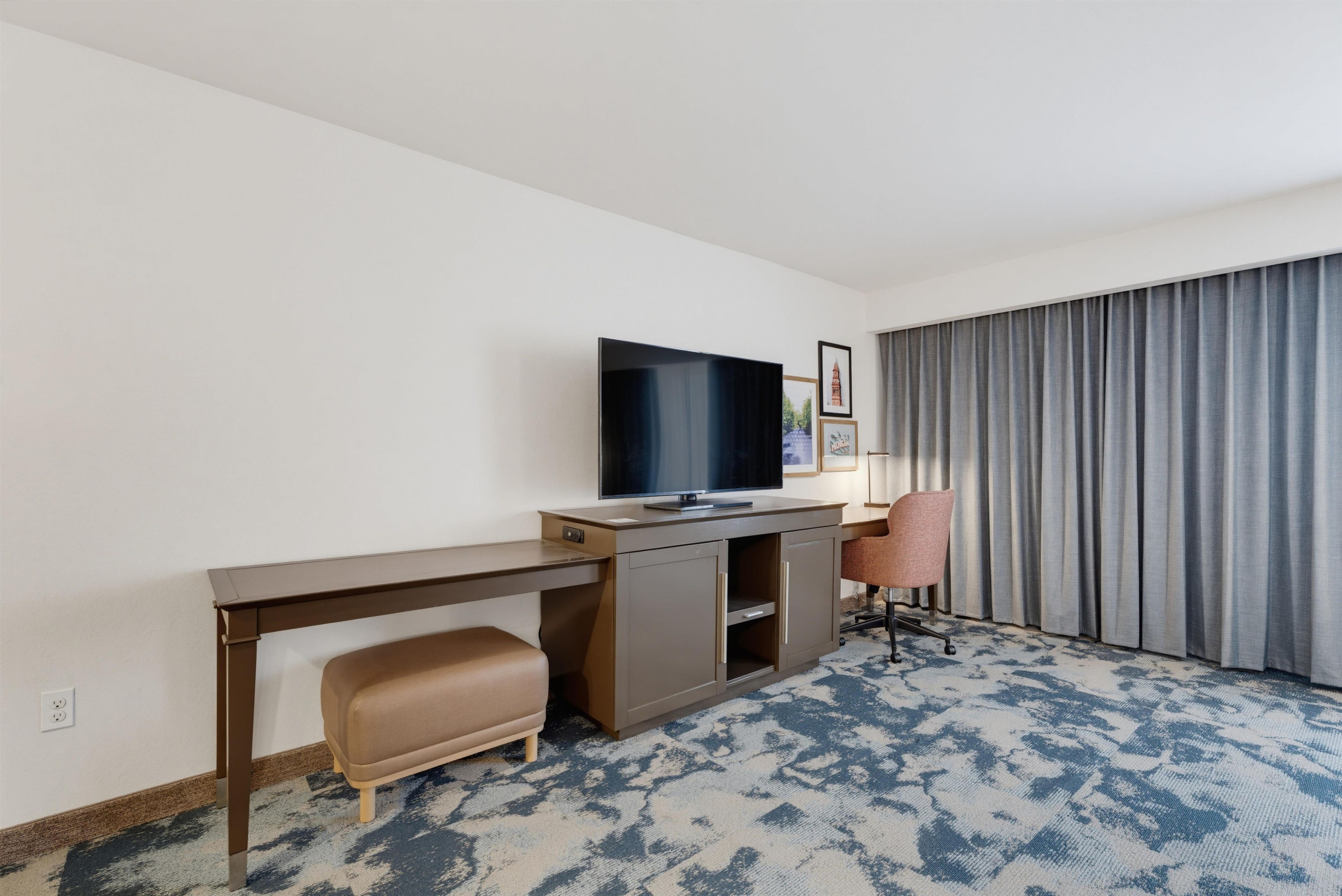 A hotel room with a flat-screen TV on a wooden desk, a cushioned bench, a single office chair, and a large window with curtains.
