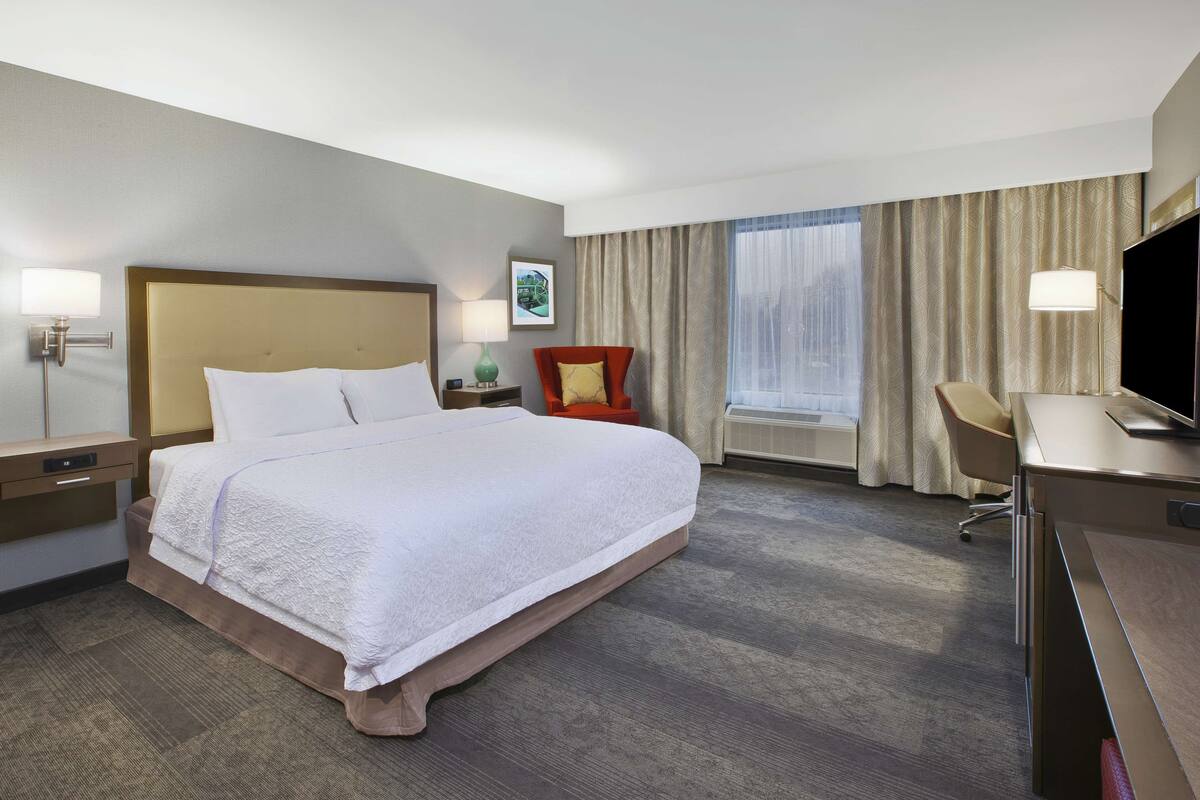 A hotel room with a large bed, a desk, a chair, a TV, and a window with curtains.