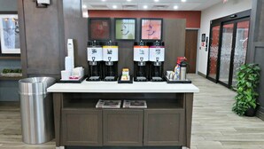 A coffee station with four dispensers, a trash can, and a small shelf with items.