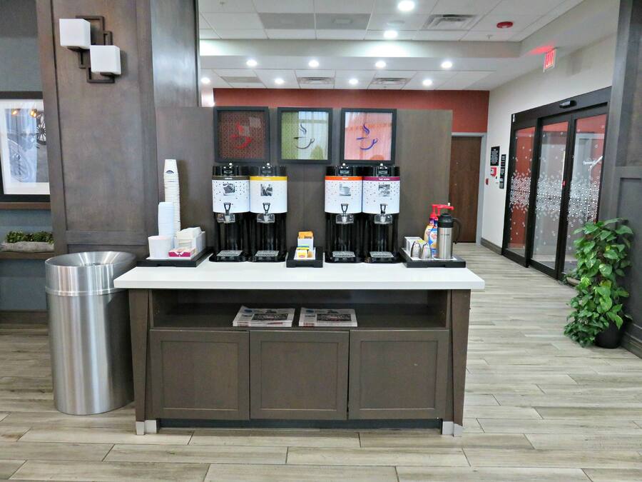A coffee station with four dispensers, a trash can, and a small shelf with items.