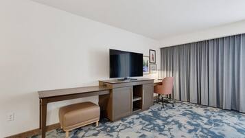 A hotel room with a flat-screen TV on a wooden desk, a cushioned bench, a single office chair, and a large window with curtains.