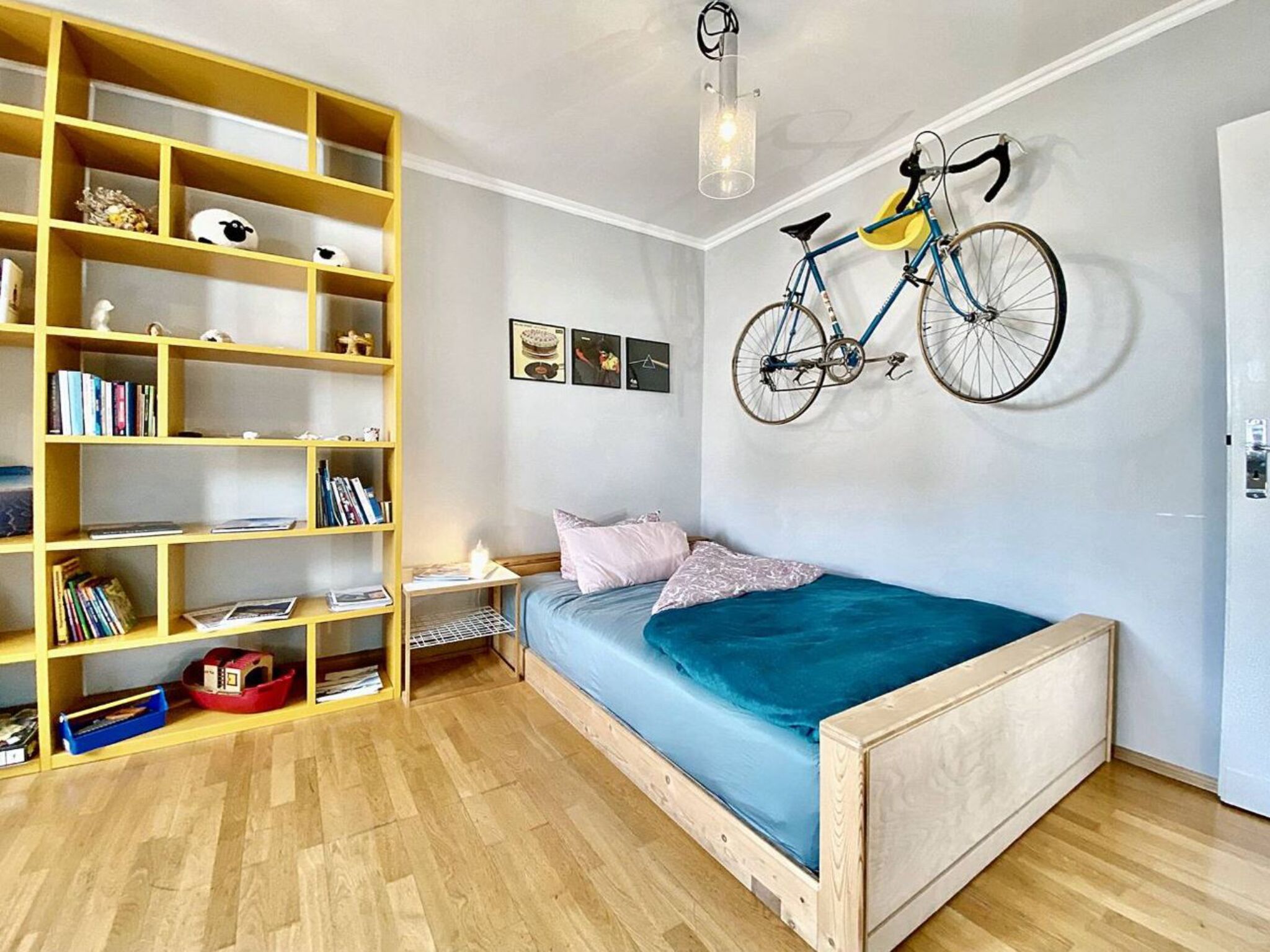 Ein Schlafzimmer mit einem hölzernen Bett, einem Bücherregal, einem an der Wand befestigten Fahrrad und einem kleinen Schreibtisch mit Stuhl.