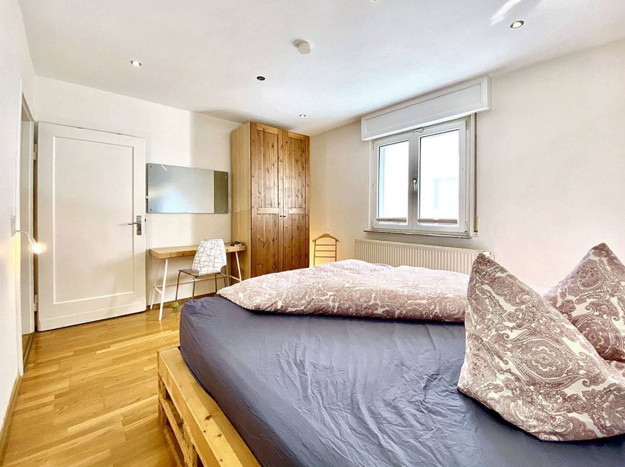 A bedroom with a bed, a desk, a wardrobe, and a window.