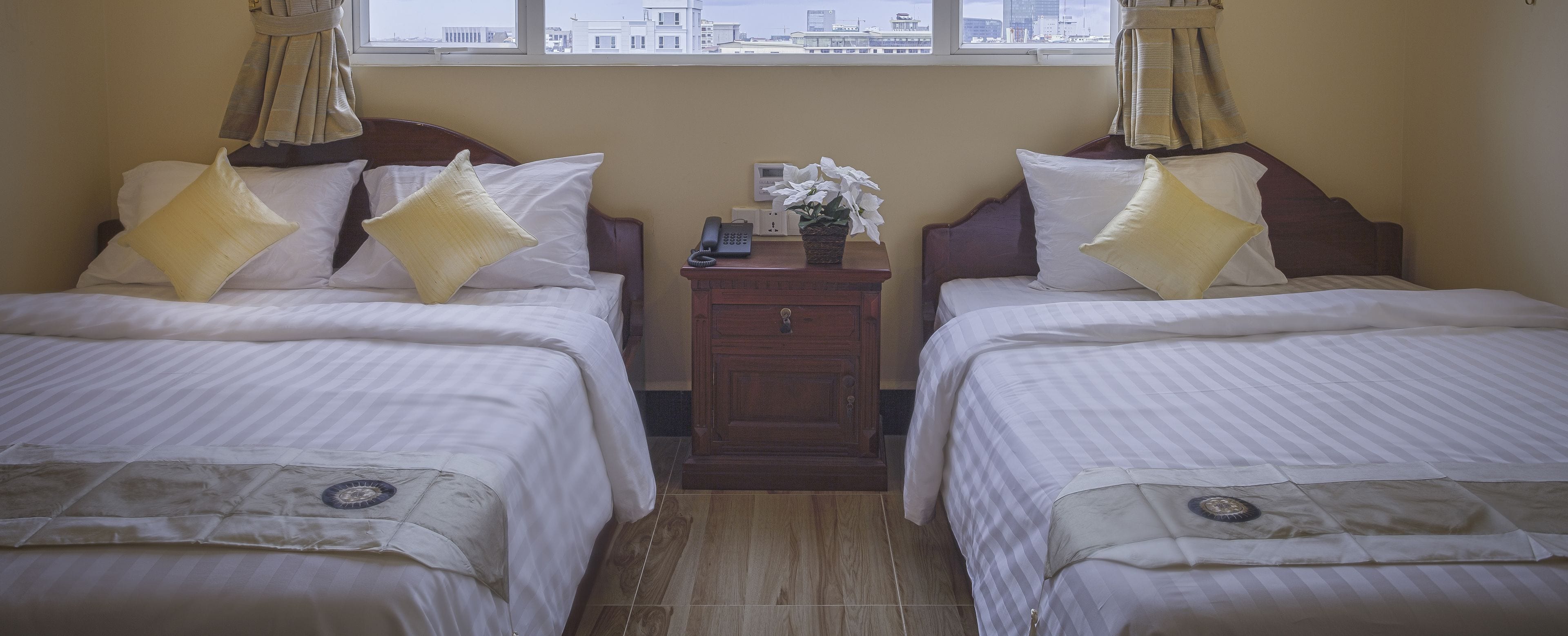 Een hotelkamer met twee bedden, een nachtkastje, een telefoon en een raam met uitzicht op de stad.