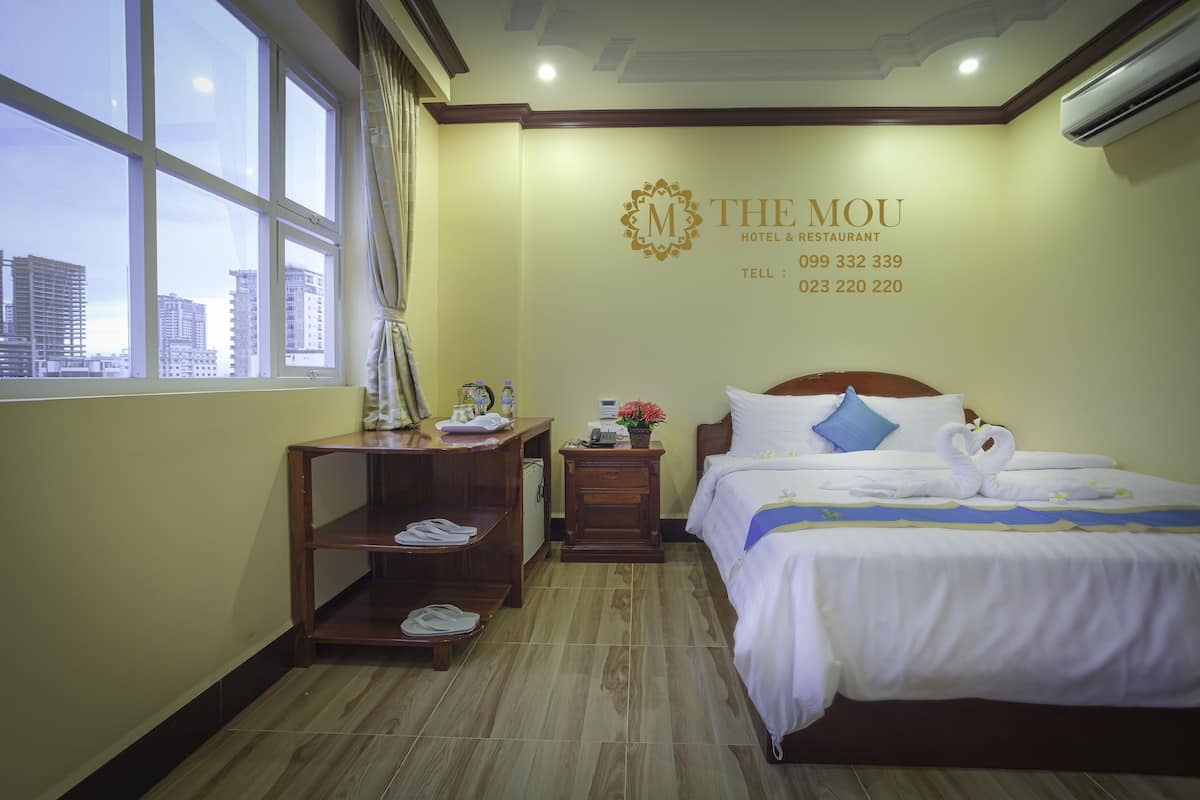 Habitación de hotel con cama, mesita de noche, estante de madera, ventana y pared con el nombre y datos de contacto del hotel.
