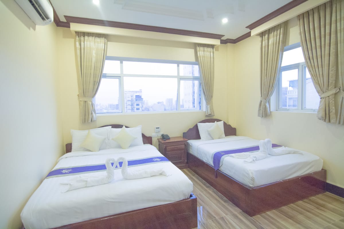 Habitación de hotel con dos camas, cada una con una toalla azul y una toalla con forma de cisne sobre la cama. Se ve una ventana con cortinas, una mesita de noche con teléfono y suelo de madera.