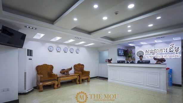 A reception area with a reception desk, two wooden chairs, a television, and a wall clock. - The Mou Hotel (Phnom Penh)