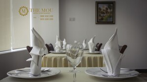 A dining table set with white napkins, a wine glass, and plates, in a room with a framed picture on the wall. - The Mou Hotel (Phnom Penh)