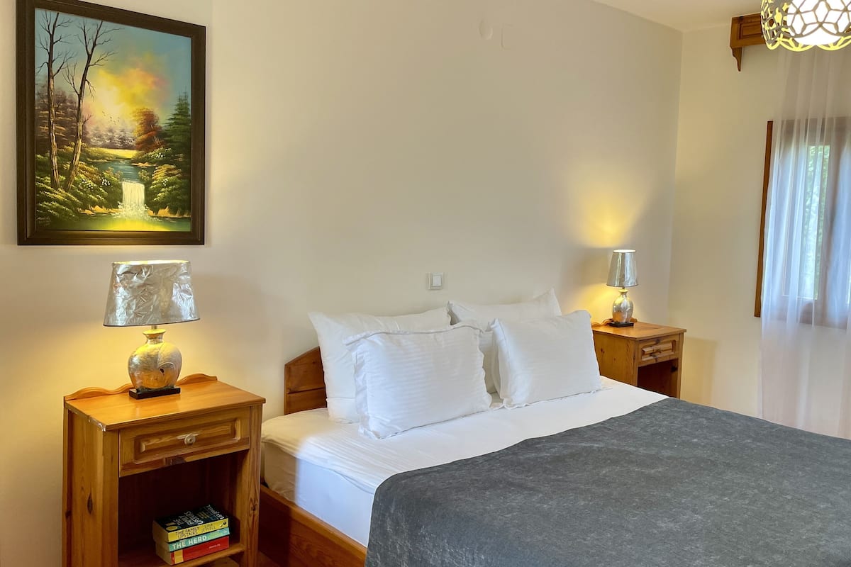 Une chambre à coucher avec un lit, deux tables de chevet, une commode, une lampe, un tableau et une fenêtre avec des rideaux.