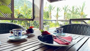 Una mesa de madera con un plato de comida, dos tazas y un vaso de agua en un balcón con vistas a la vegetación.