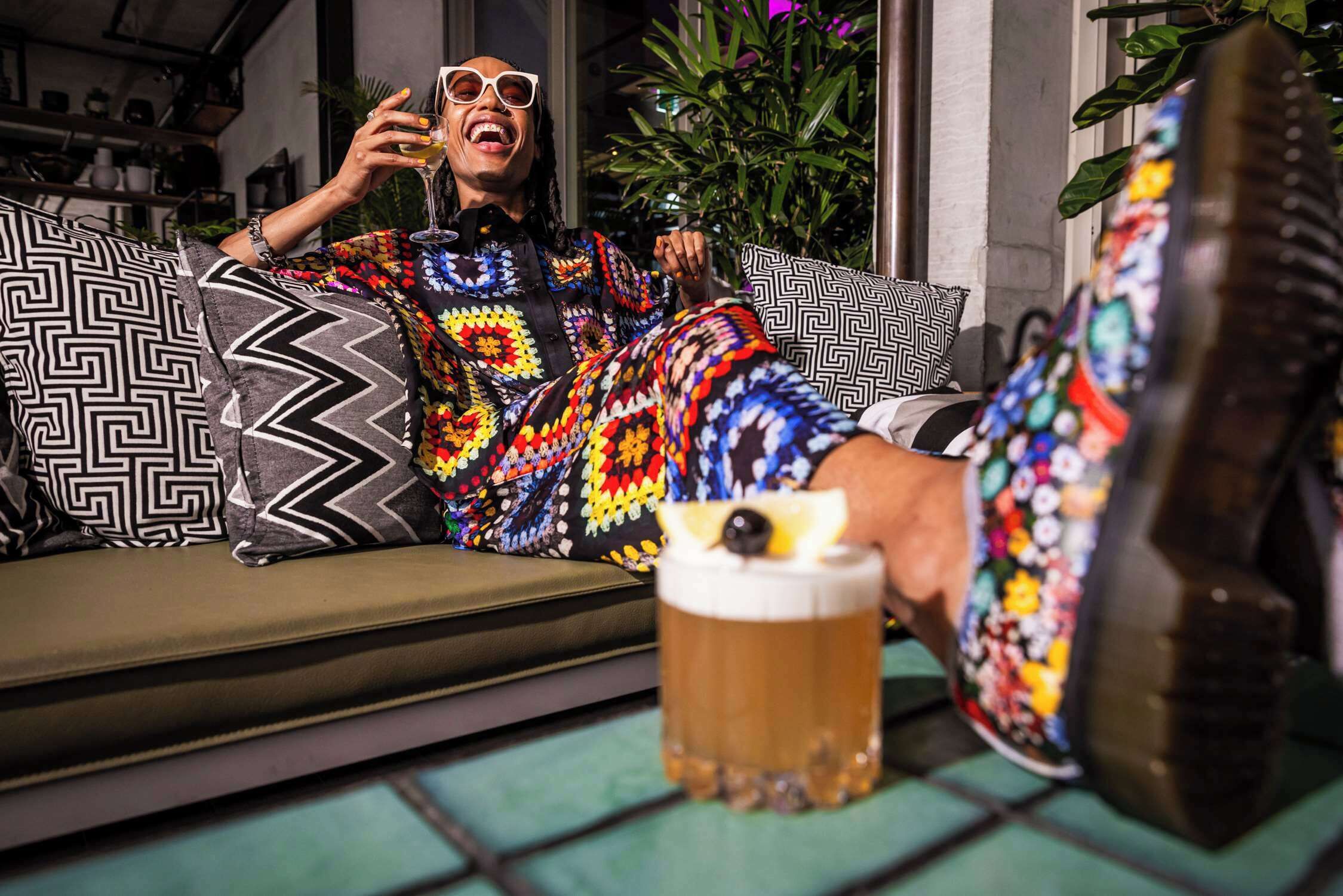 Una persona sentada en un sofá con un cojín estampado, sosteniendo una jarra de cerveza y vistiendo ropa colorida.