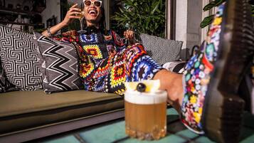 Una persona sentada en un sofá con un cojín estampado, sosteniendo una jarra de cerveza y vistiendo ropa colorida.