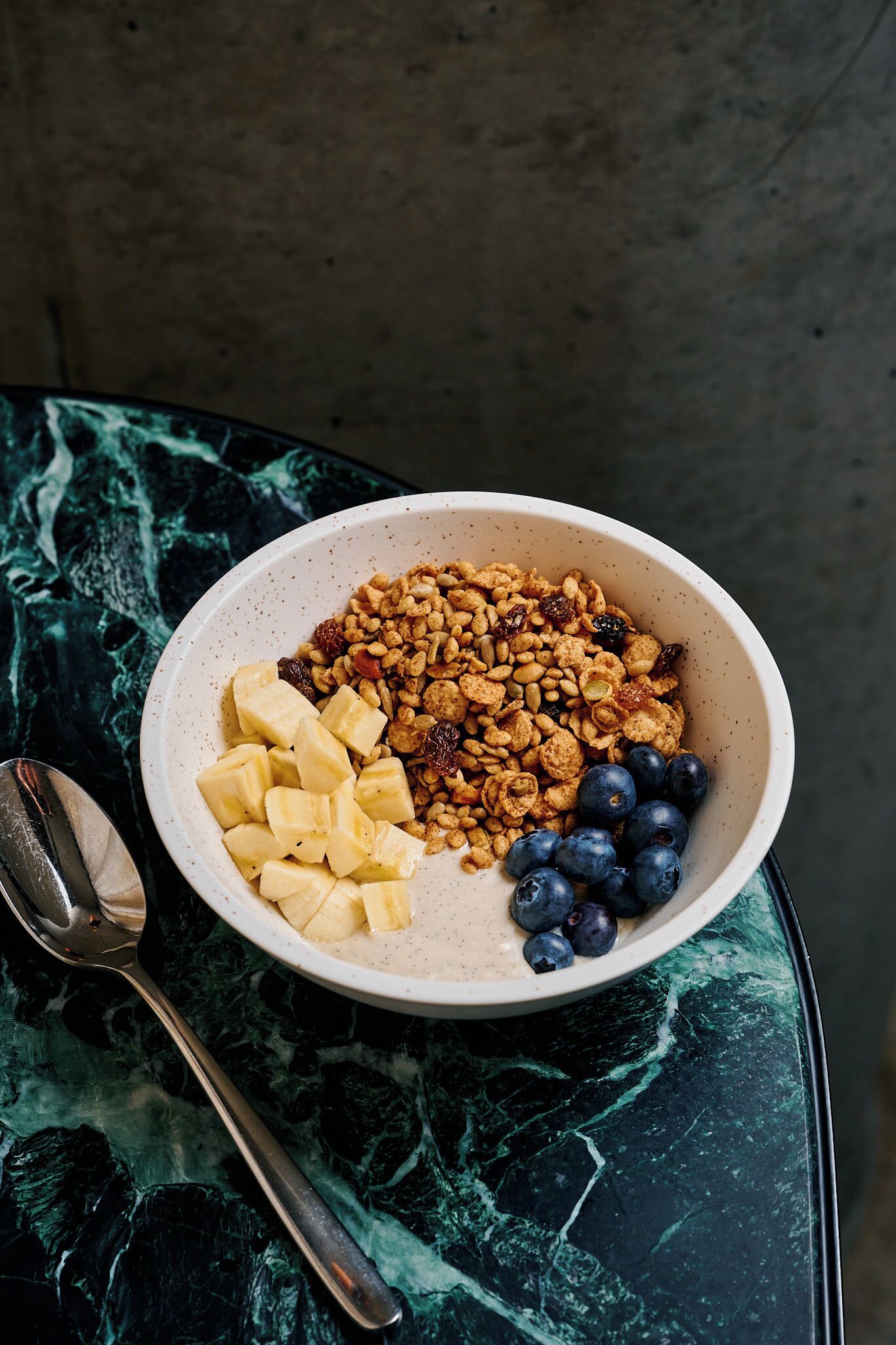 A bowl of granola with bananas and blueberries on a marble table.
