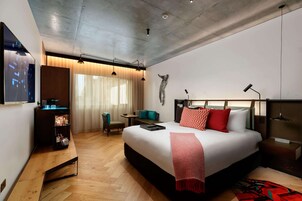A modern hotel room with a large bed, a desk, a chair, and a TV on the wall.