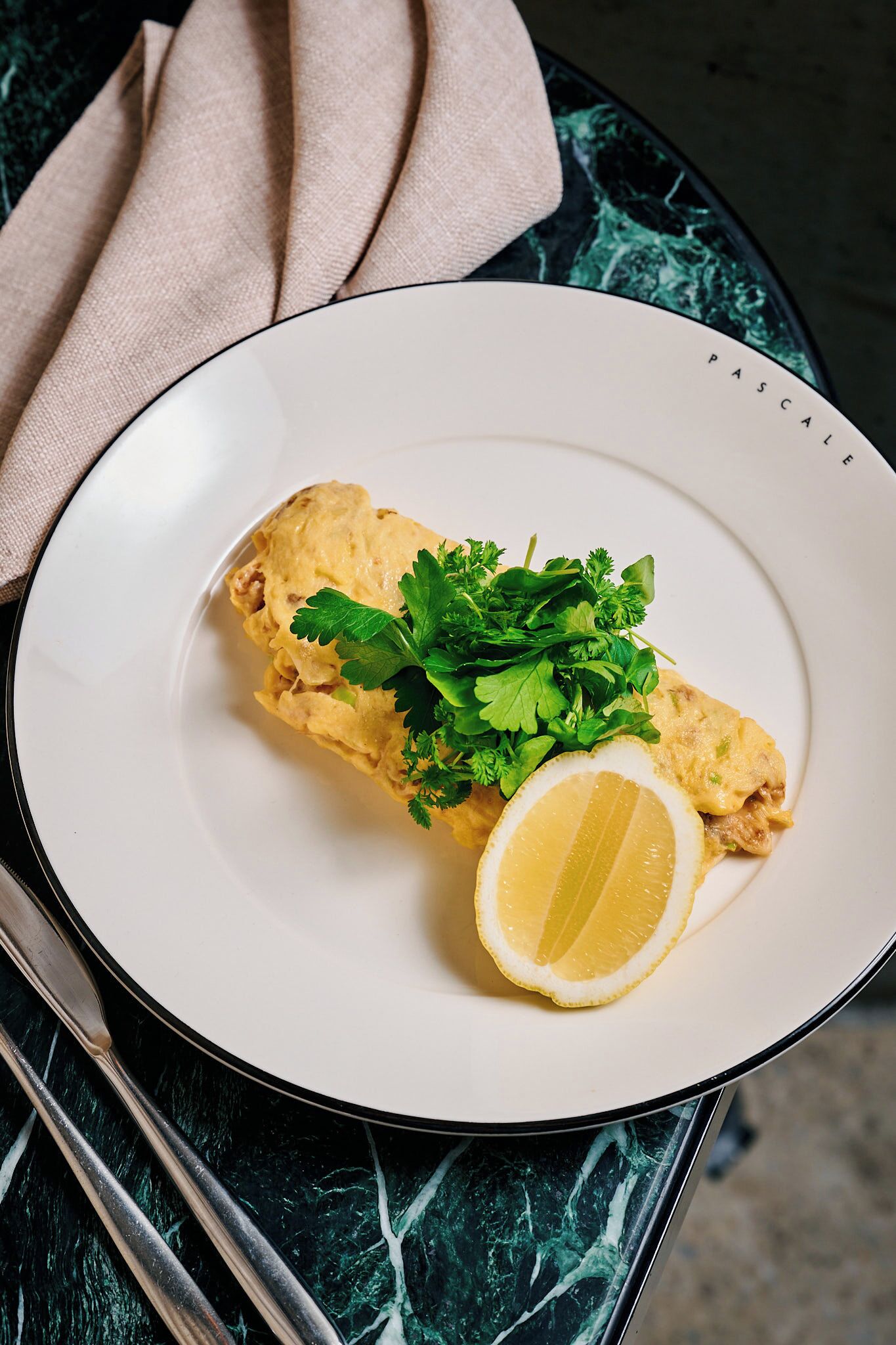 A plate with a rolled omelette, parsley, and a lemon slice on a white plate with a black rim.