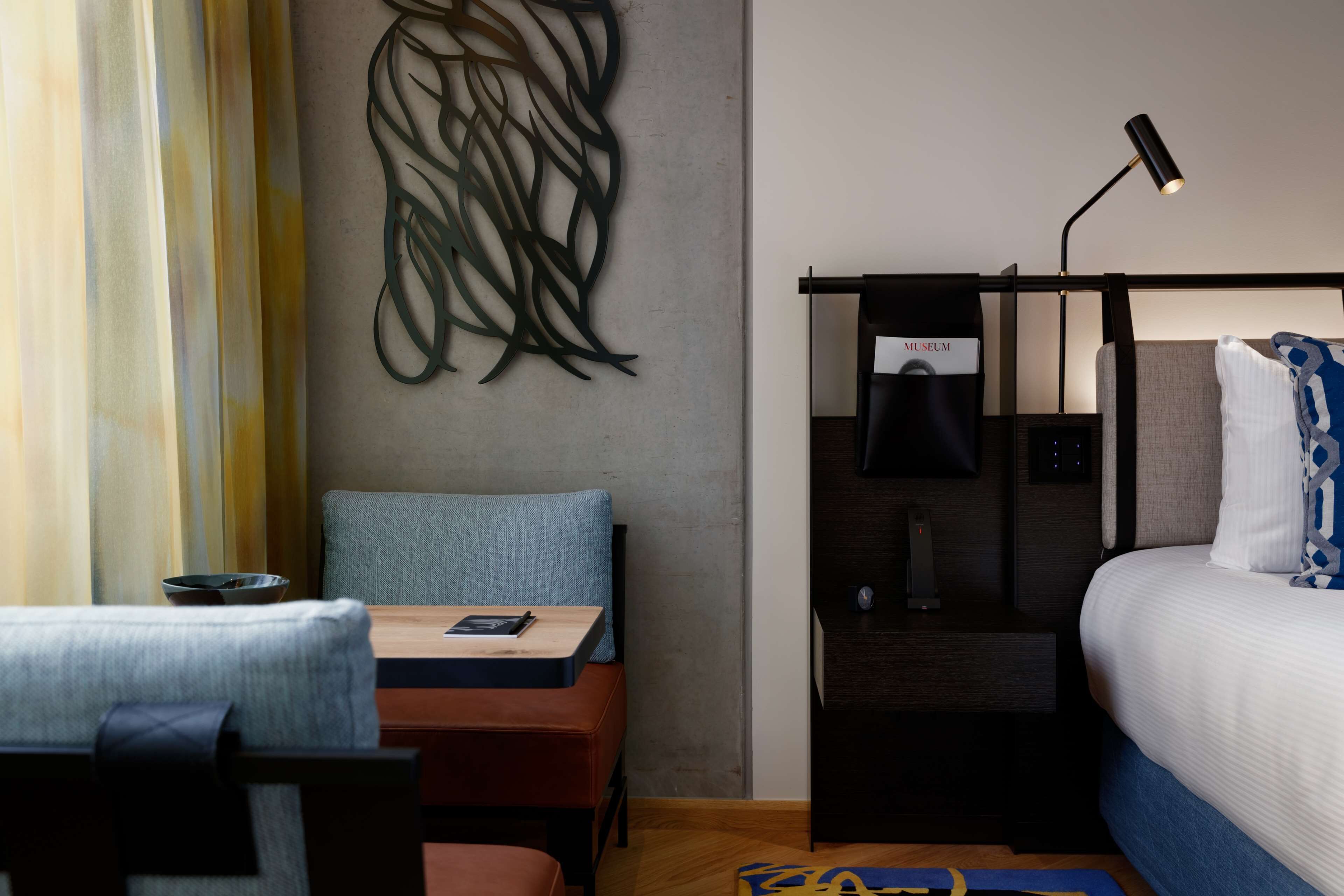 Una habitación de hotel moderna con cama, escritorio con silla, mesita de noche y una obra de arte colgada en la pared.