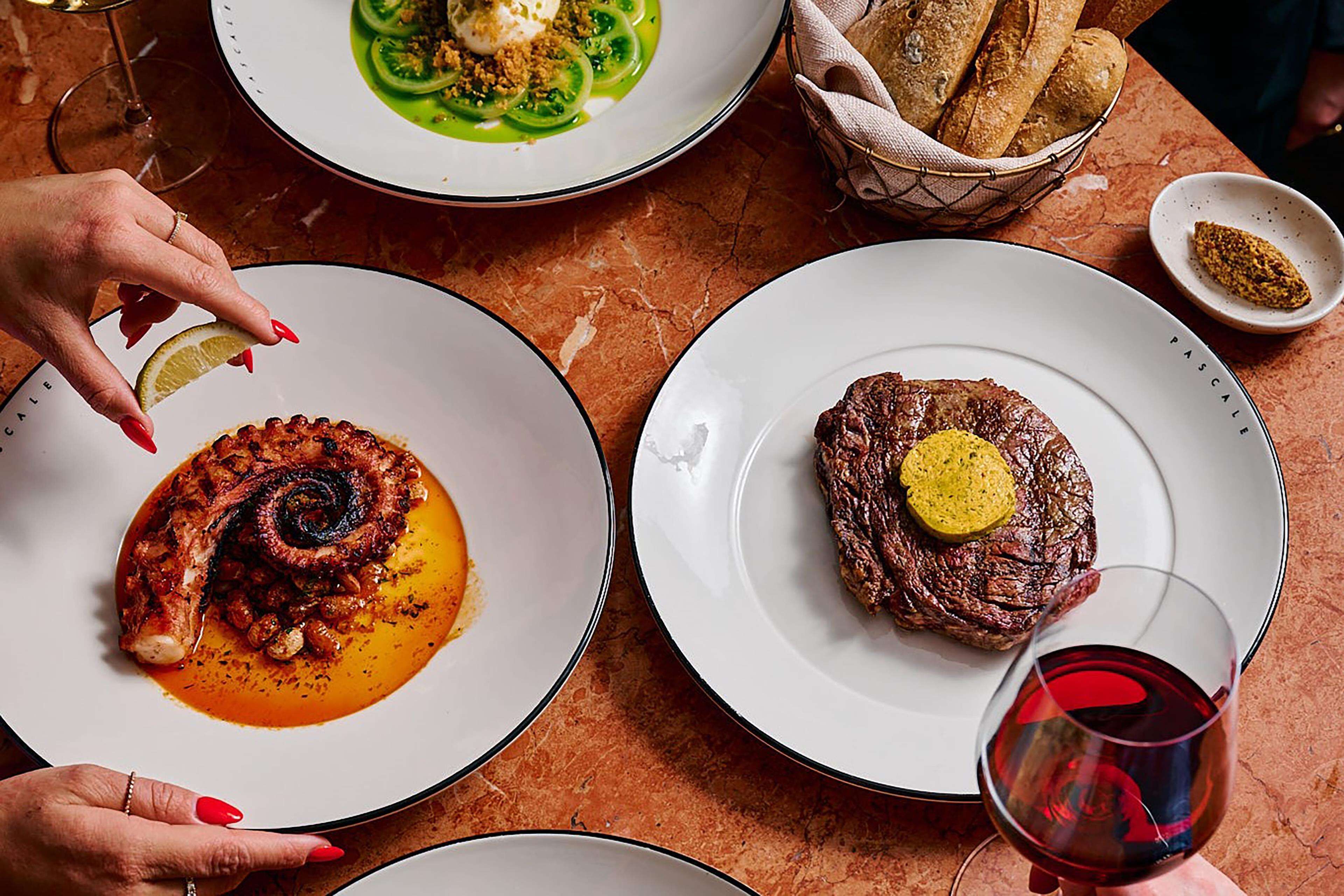 Una mesa con platos de comida, una cesta de pan y una copa de vino tinto.