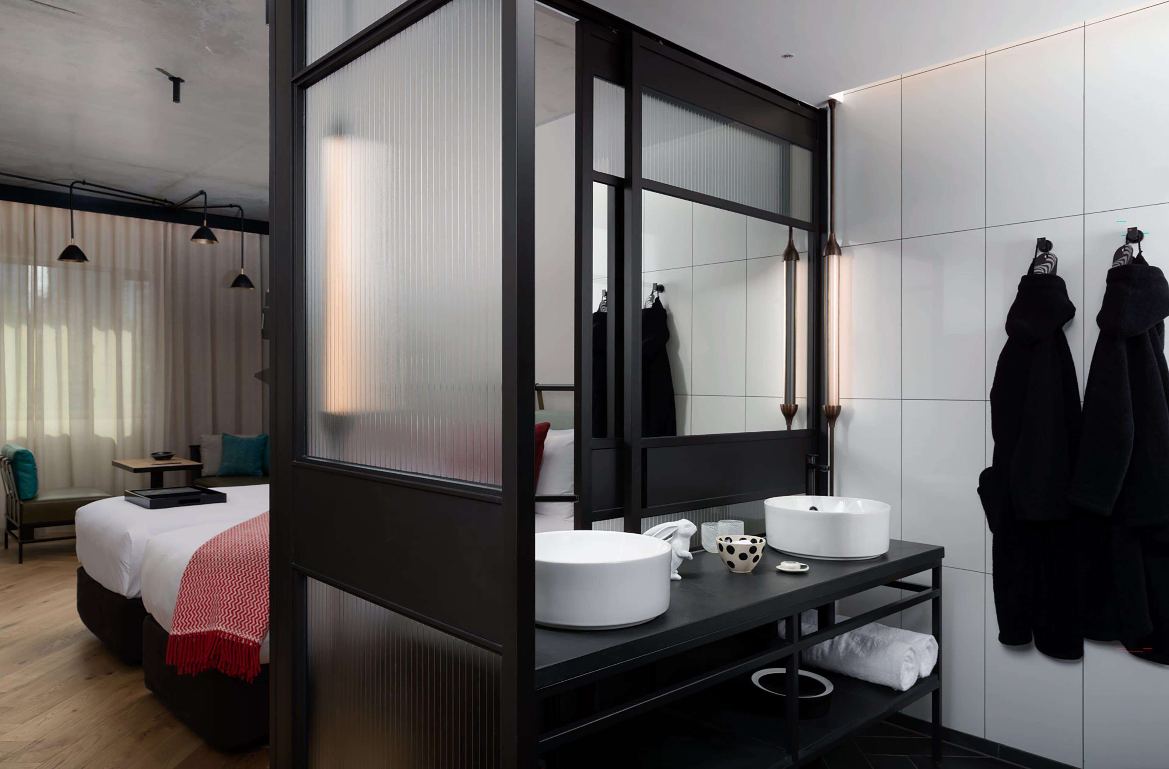 A modern hotel room with a large mirror, two white round sinks, a black vanity, a bed with white linens, a red throw blanket, a small table, and a chair.