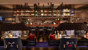 Zona de bar con estante lleno de botellas, dos sillas con respaldos decorativos y una barra con espejo.