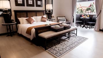 A hotel room with a large bed, bedside lamps, a bench, a small table with fruit and drinks, and a view of the outside.