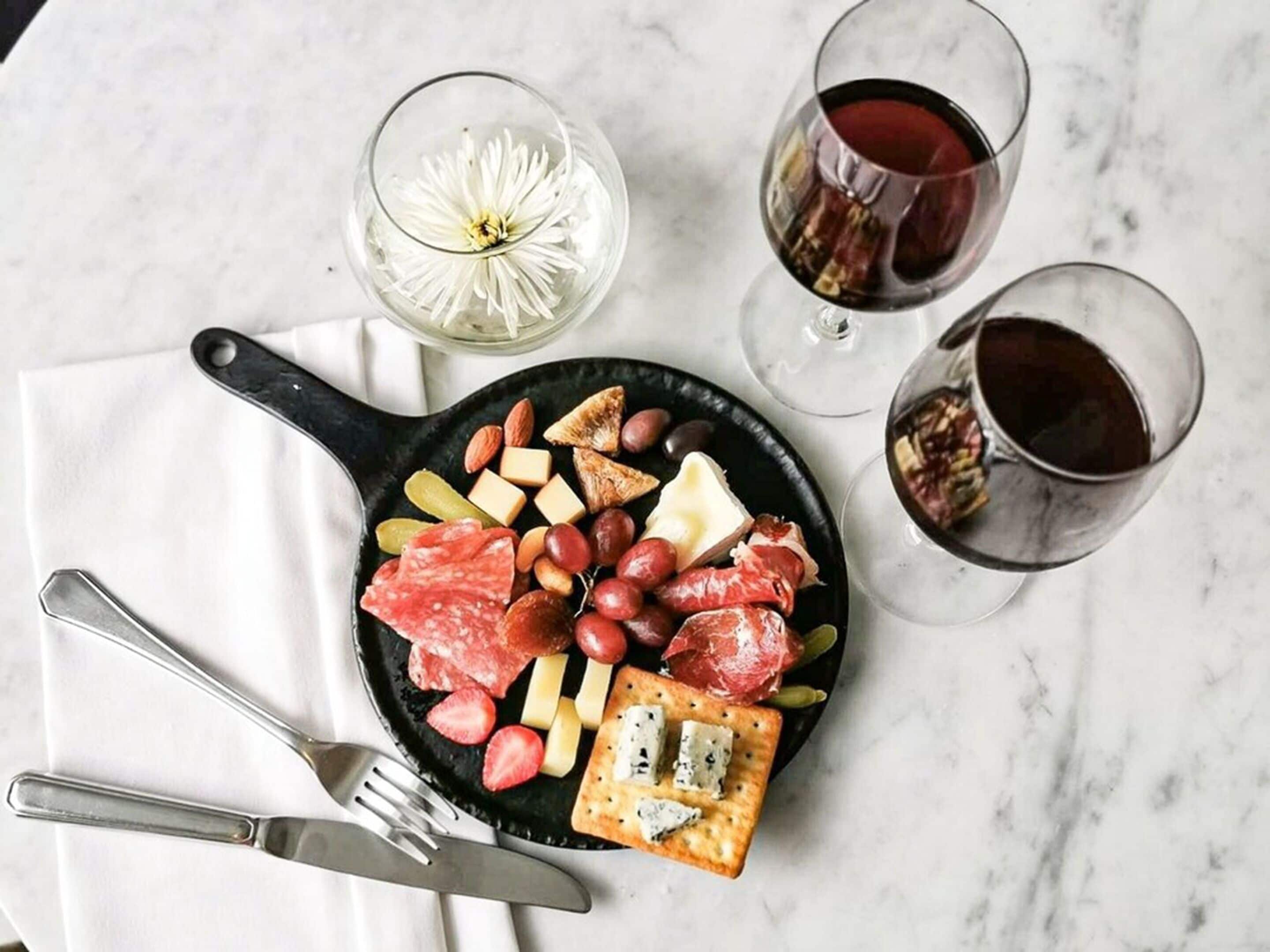 A charcuterie board with various meats, cheeses, and crackers, accompanied by two glasses of red wine and a flower in a glass.