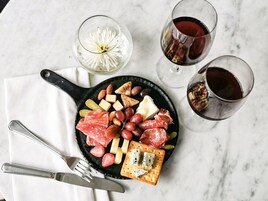 A charcuterie board with various meats, cheeses, and crackers, accompanied by two glasses of red wine and a flower in a glass.