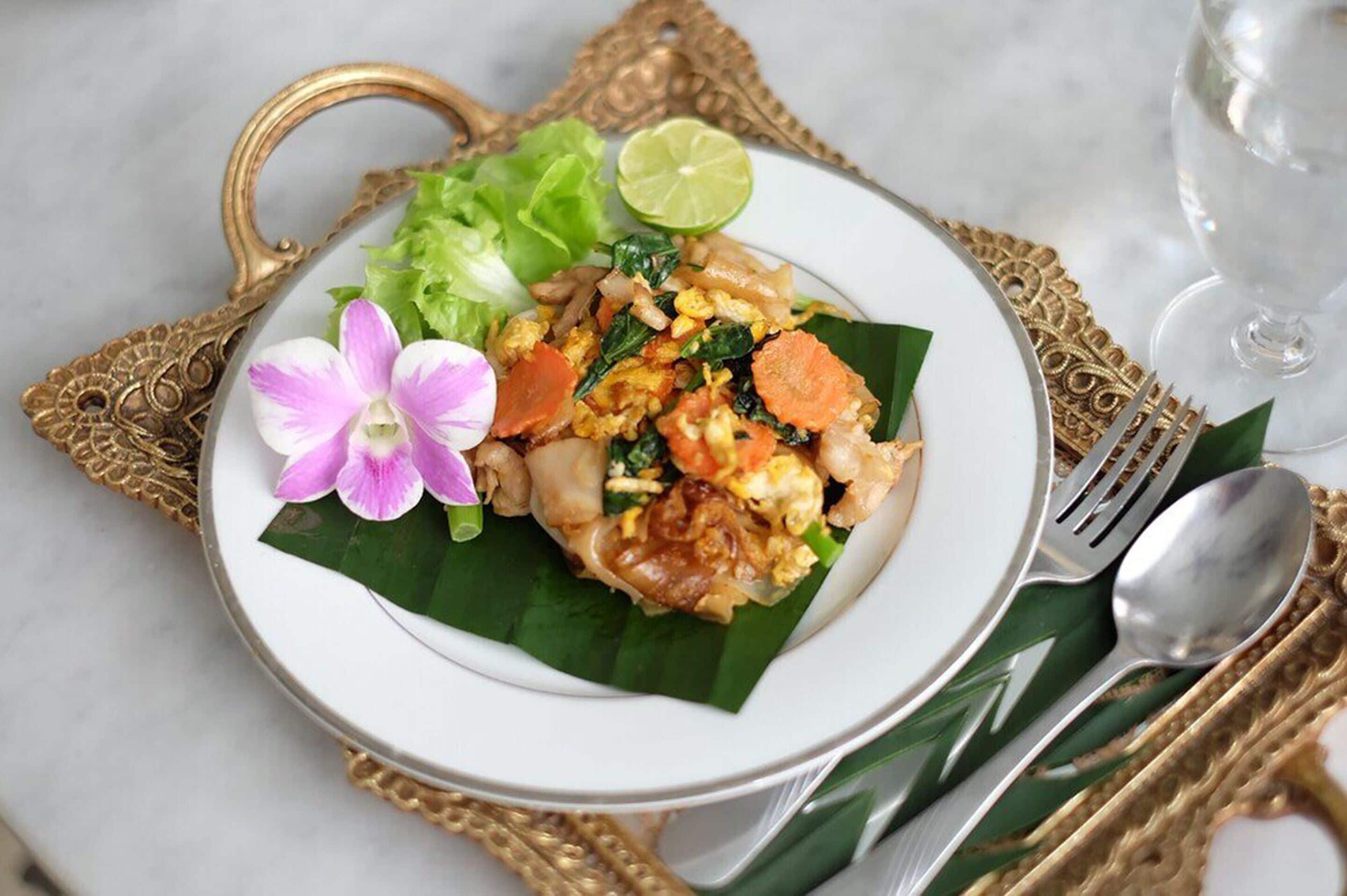 A plate of Pad Thai with vegetables and lime, served on a white plate with a gold handle.