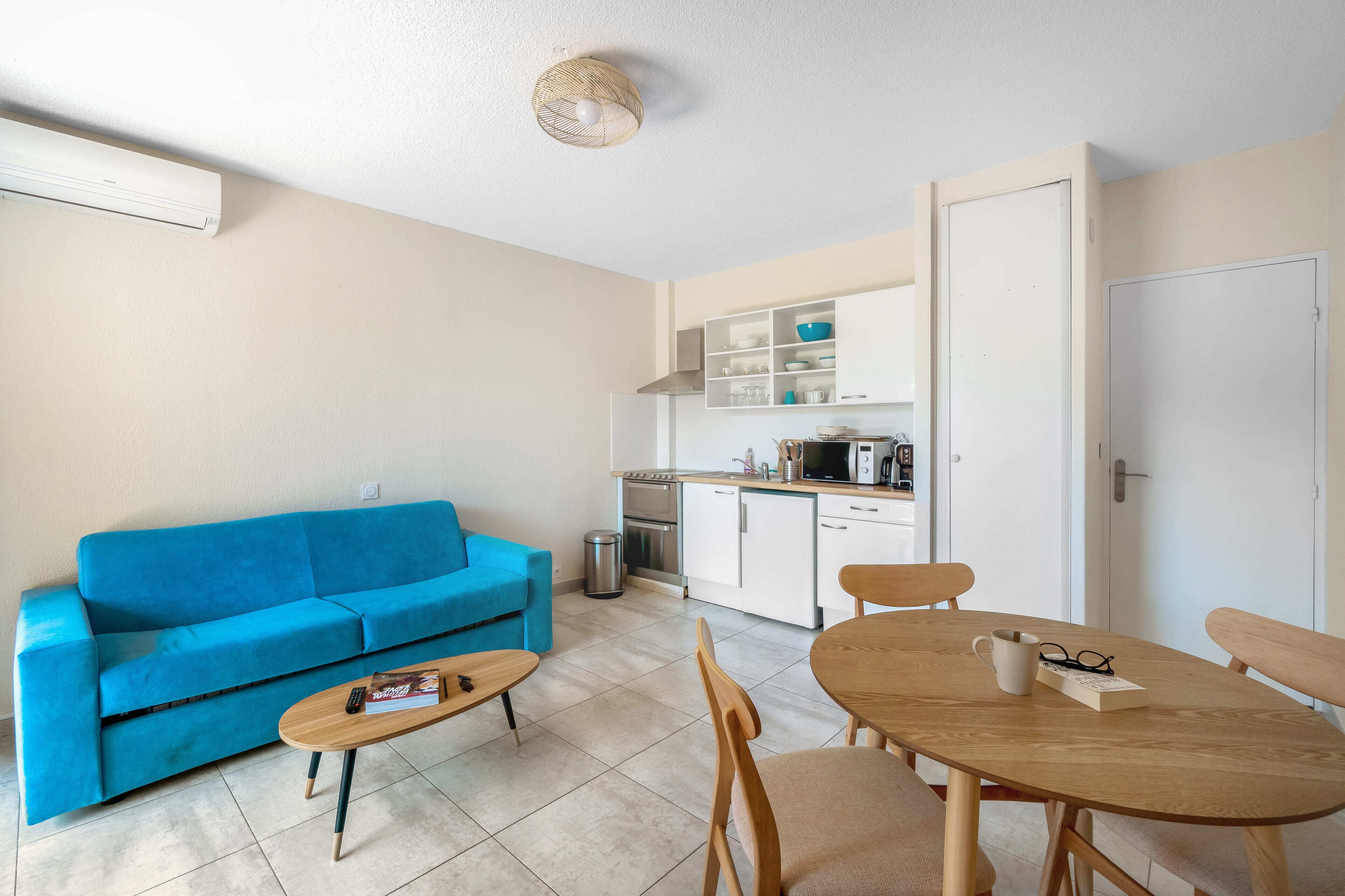 Un espacio habitable compacto con un sofá azul, una mesa de centro de madera, una mesa de comedor con sillas, una kitchenette y una puerta blanca.